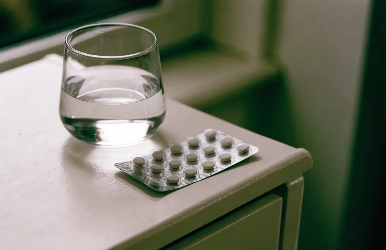 An unlabelled blister pack of tablets beside a glass of water on a nightstand